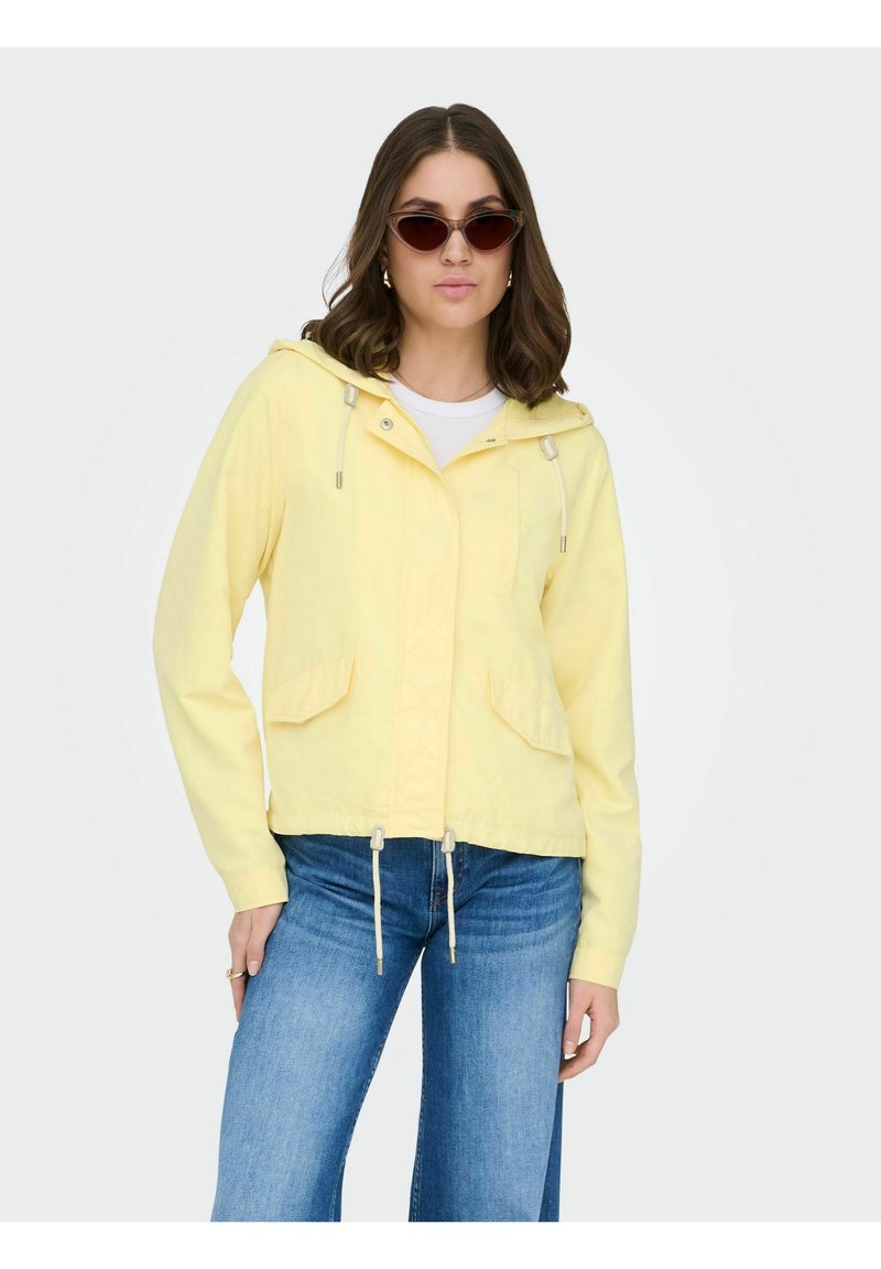 Femme portant une veste jaune à capuche avec cordons, un jean bleu et des lunettes de soleil œil de chat, debout devant un fond uni.