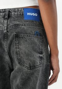 Černé džíny z denimu s vybledlou texturou. Mají modrou koženou etiketu s nápisem "HUGO" a modrý akcent poblíž zadní kapsy.