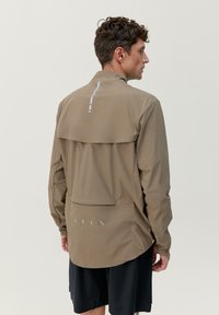 Ljust brunt nylonjacka med långa ärmar, en bakre ventil och reflekterande detaljer. Har en logotyp tryckt på nedre delen av ryggen.