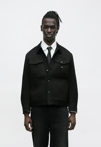 Čierna denimová bunda s prednými gombíkmi, dvoma náprsníkovými vreckami a kontrastným golierom. Nosená cez bielu košeľu a čiernu kravatu, v kombinácii s tmavými džínsami.