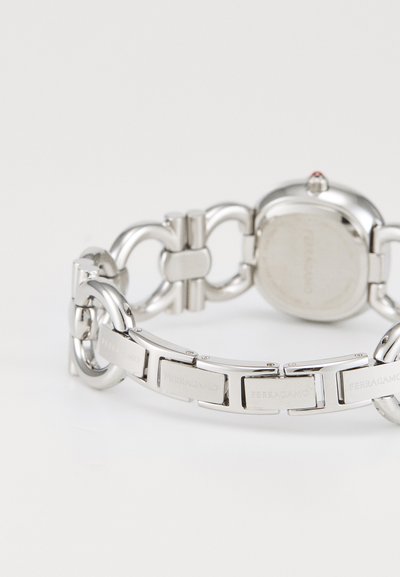 Reloj de metal plateado con diseño de pulsera enlazada, con caja circular, nombre de la marca grabado y un detalle rojo en la corona.