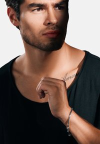 Svart skjorta på en person med en tatuering på axeln. Personen har ett silverarmband och har ett neutralt uttryck, belyst från en sida.