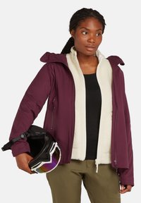 Chaqueta de esquí burdeos con forro de felpa blanca, cremalleras y bolsillos laterales. El modelo sostiene un casco negro con visera multicolor y lleva pantalones en color oliva.