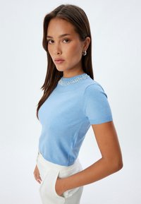 Maglione a maniche corte di un azzurro chiaro con un design testurizzato e dettagli decorativi in perline attorno al colletto, abbinato a pantaloni bianchi a vita alta.