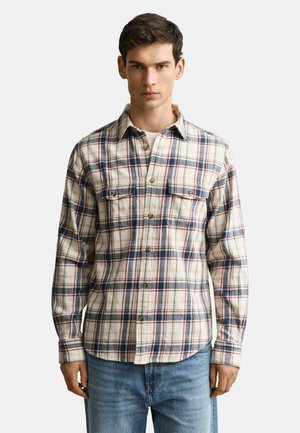 Jeune homme portant une chemise à carreaux crème, bleu marine et rouge avec deux poches poitrine et un jean bleu clair, debout devant un fond blanc.