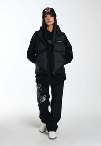 Schwarze Puffer-Weste über einem schwarzen Hoodie, kombiniert mit schwarzen Hosen, die mit buntem Fischstickereien verziert sind, und weißen Sneakers. Schwarze Kappe mit Grafik.