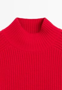 Detailní záběr na červený pletený svetr s vysokým, širokým rolákem na jednoduchém bílém pozadí.