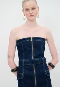 Denim-tyylinen olkaimeton mekko, jossa on etuvetoketju, vyötärölle tarkoitettu vyö ja kaksi etutaskua. Tumma sininen väri, sileä tekstuuri ja jämäkkä muotoilu.