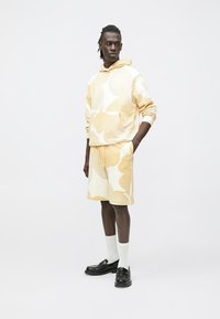 Conjunto de sweatshirt com capuz e calções em amarelo pastel com padrões abstratos em creme; material de mistura de algodão, com bolsos laterais, usado com meias brancas e sapatos pretos.