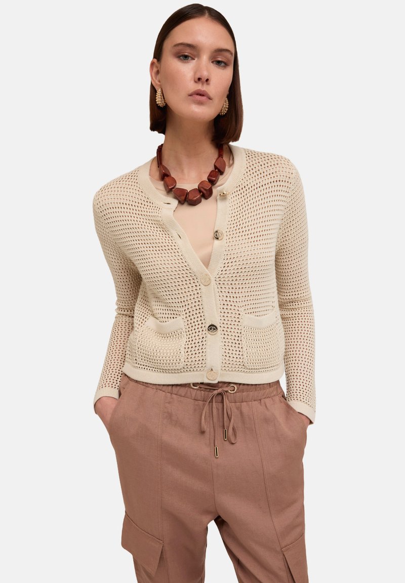 Beige, avointa neuletta oleva neuletakki, jossa on pyöreä kaula-aukko, kaksi etutaskua ja nappikiinnitys. Yhdistetty vaaleanruskeisiin, naruvaljailla varustettuihin housuihin.