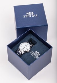 Sidabrinis metalinis laikrodis su baltu ciferblatu, datos rodymu ir juodais akcentais, pateiktas tamsiai mėlynoje dėžutėje su aksominiu vidumi.