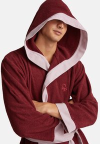 Burgundská koupací pláštěnka s kapucí v měkké, strukturované tkanině. Má světle růžové lemování a vyšívané logo na prsou.
