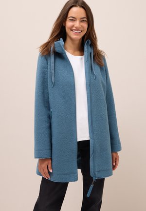Femme souriante portant une veste zippée bleue texturée sur un haut blanc et un pantalon noir devant un fond beige uni.
