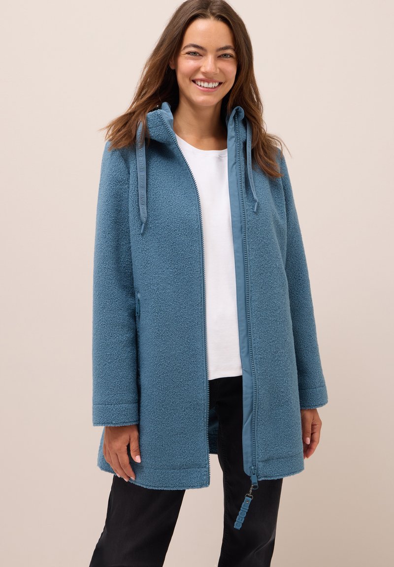 Femme souriante portant une veste zippée bleue texturée sur un haut blanc et un pantalon noir devant un fond beige uni.
