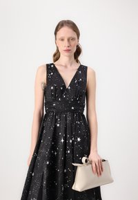 Black dress with white star pattern, deep V-neckline, fitted waist, and full skirt; holding a beige structured handbag.