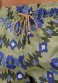 Shorts de bain multicolores en vert, présentant un motif géométrique bleu, noir et blanc. Taille élastique avec un cordon de serrage beige.