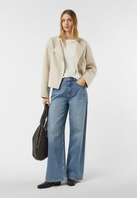 Beżowa krótka kurtka, biała koszulka i szerokie niebieskie dżinsy z denimu. Modelka trzyma czarną teksturowaną torbę. Całość dopełniają czarne baleriny.