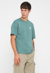 Turquoise cotton T-shirt with short sleeves and a round neck, featuring a small white balloon graphic on the left chest.