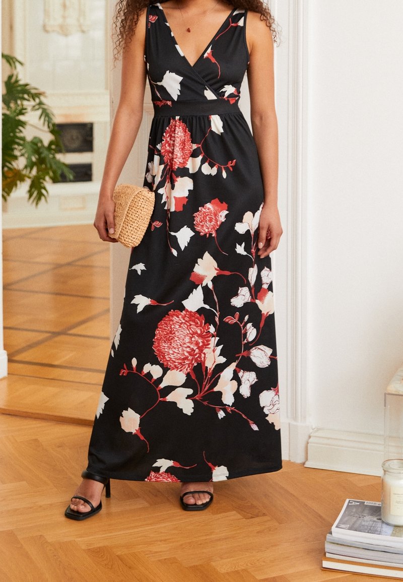 Femme portant une robe longue noire à fleurs rouges et blanches, tenant une pochette tissée beige, debout sur un sol en bois à l'intérieur.