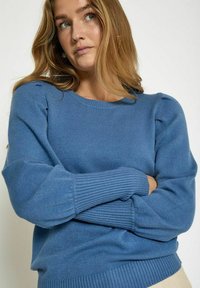 Pull en tricot bleu avec un col rond, des épaules bouffantes, et des poignets et un ourlet côtelés. Texture douce visible dans le tissu.
