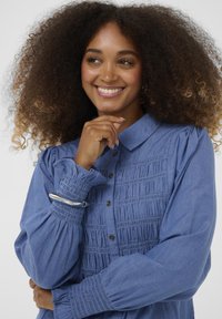 Sininen chambray-paita, jossa on smokkaukset, nappisuljenta, kauluksellinen kaula ja kerätyt hihat. Hopeinen korulenkki ranteessa.