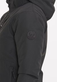 Schwarze wetterfeste Jacke mit zipbarem Taschen, verstellbarem Kapuzen und einem runden Logo-Patch auf dem Oberarm. Glatte Textur.