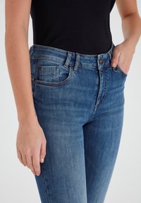 Augstas jostas zilie džinsi ar izbalējušu tekstūru, piecu kabatu dizains un metāla pogas aizdare jostasvietā.