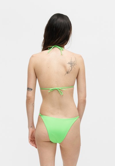 Γυναίκα με την πλάτη, φοράει lime πράσινο μπικίνι με δεμένα λουριά, μακριά σκούρα μαλλιά και εμφανή τατουάζ στην άνω πλάτη και στο αριστερό χέρι.