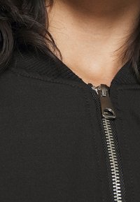 Černá zipová bunda s kovovým zipem, texturovanou látkou a žebrovaným límcem. Detailní pohled na oblast výstřihu s viditelnými švy.