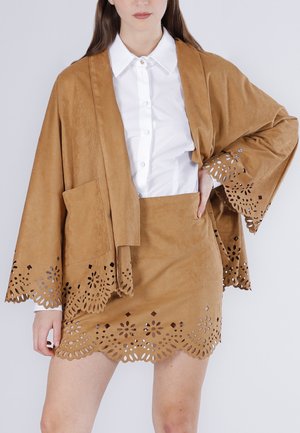 Mujer con falda de ante color beige y chaqueta estilo kimono a juego con bordes florales cortados con láser, combinada con una camisa blanca abotonada.