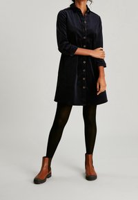 Granatowa sukienka koszulowa z sztruksu z długimi rękawami, z przodu zapinana na guziki i o rozkloszowanym kroju. W połączeniu z brązowymi botkami i czarnymi rajstopami.