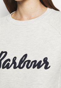 Světle šedá mikina s kulatým výstřihem, s vyšívaným logem "Barbour" v námořnicky modré barvě napříč přední částí. Měkká textura a raglánové rukávy.