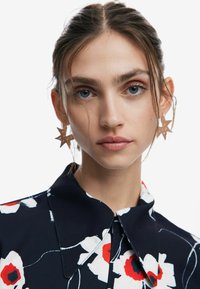 Blusa floreale navy con motivi bianchi e rossi, caratterizzata da un ampio colletto e chiusura a bottoni; il modello indossa orecchini a forma di stella.