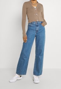 Kvinna i en beige knäppt kofta, hög midja blå vida jeans, vita sneakers och ett silverkedjehalsband.