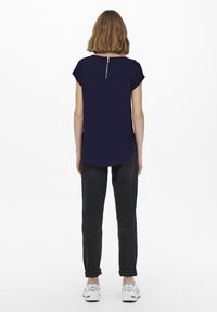 Tmavě modré tričko s krátkým rukávem a kulatým výstřihem, se zlatým zipem na zádech, v kombinaci s tmavými džíny a bílými teniskami. Hladká textura.