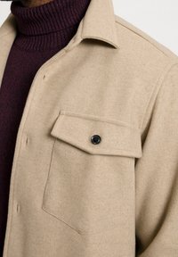 Beżowa koszula-kurtka z mieszanki wełny, z kołnierzem, zapięciem na guziki z przodu oraz pojedynczą kieszenią na piersi z czarnym detalem w postaci guzika.
