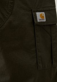 Ciemnozielony materiał o fakturze kratki, z boczną kieszenią i naszywką z logo Carhartt przyszytą do kieszeni.