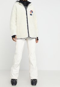 Vit fuskpälsjacka med huva, marinblå detaljer och en logotypplåster; matchad med vita skidbyxor och svarta skidstövlar.