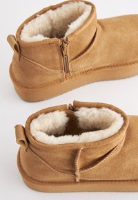 Tanfarvede ankelstøvler i ruskind med foret af pels, lynlåslukning og detalje med trækstrop. Blød tekstur med flad sål for komfort.