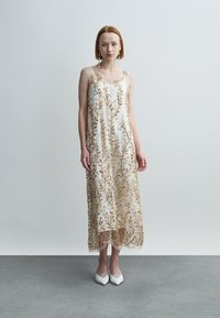 Λευκό, μήκους έως το γόνατο φόρεμα με χρυσά κεντημένα σχέδια φύλλων. Απαλό, διάφανο ύφασμα. Το μοντέλο φοράει λευκά παπούτσια. Απλές τιράντες και ρέον σιλουέτα.