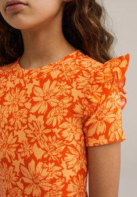 Oranžinis juostuotas viršus su gėlių raštu, trumpomis volanomis ir apvaliu kaklu. Dizainas sujungia tamsesnius ir šviesesnius oranžinius atspalvius.