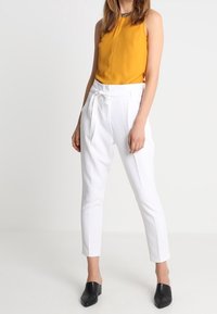 Yellow sleeveless top paired with white trousers featuring a tie waist. Model wears black mules, with a smooth fabric finish overall.