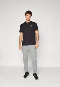 Mladi moški stoji oblečen v črno majico Puma, sive trenirke Puma in črni športni copati na enobarvnem svetlem ozadju.