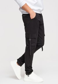 Vue de profil d'une personne portant un pantalon cargo noir avec des poches et des cordons, associé à des baskets blanches et un sweat-shirt gris clair.