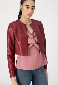 Giacca cropped in pelle bordeaux con scollatura rotonda e maniche lunghe, dotata di una cerniera d'oro. Indossata sopra un top a strati di colore rosa chiaro.
