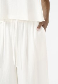 Weißes, strukturiertes Stoffoberteil und weit geschnittene Hose. Das Oberteil fällt locker und lässig, die Hose hat einen Kordelzug und seitliche Taschen.