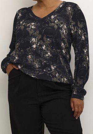 Frau trägt schwarzen Langarmoberteil mit abstraktem weißem und grünem Muster, kombiniert mit schwarzer Hose, steht mit einer Hand in der Tasche.