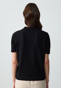 Polo sweater nero a maniche corte con una texture liscia, polsini a maglia e un colletto discreto. La vista posteriore mostra un design aderente e linee pulite.