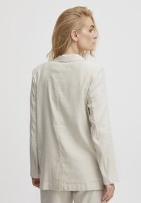 Hellgrauer Leinenblazer mit taillierter Silhouette, scharfen Revers und seitlichen Schlitzen. Die glatte Textur und minimalen Designdetails unterstreichen die Einfachheit.