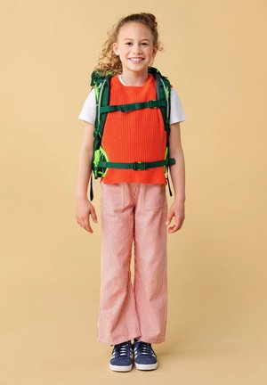 Young girl with curly hair smiling, wearing an orange sweater vest, striped pants, navy sneakers, and a green backpack with straps fastened.